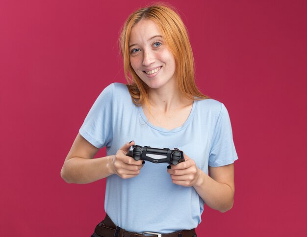 Sonriente joven pelirroja pelirroja con pecas con controlador de juego en rosa