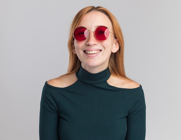 Sonriente joven pelirroja pelirroja con pecas en gafas de sol mirando a cámara en blanco