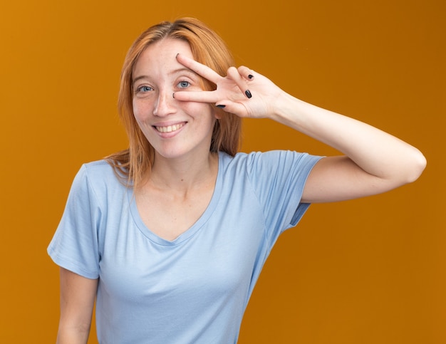 Sonriente joven pelirroja pelirroja con pecas gestos signo de victoria mirando a la cámara a través de los dedos en naranja