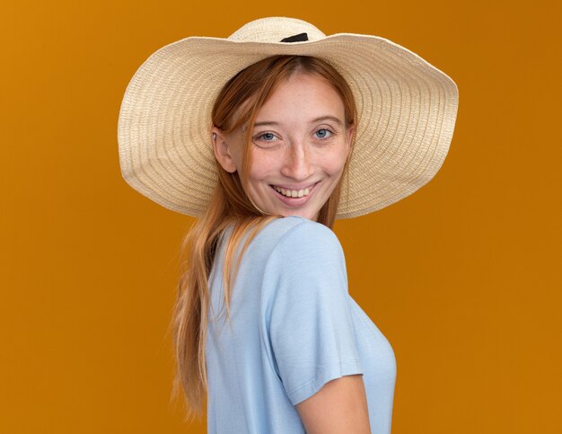 Sonriente joven pelirroja pelirroja con pecas vistiendo sombrero de playa de pie hacia los lados en naranja