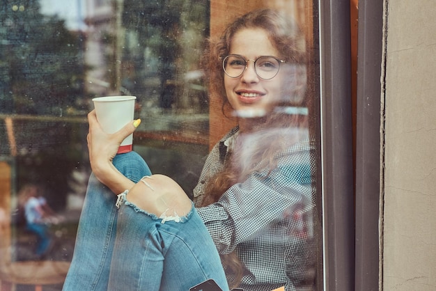 Sonriente joven pelirroja rizada con ropa informal y gafas sentada en un alféizar con un café para llevar.