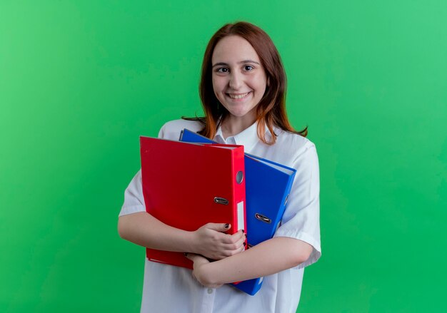 Sonriente joven pelirroja sosteniendo carpetas aisladas en verde