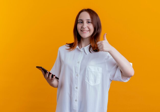 Sonriente joven pelirroja sosteniendo el teléfono y su pulgar hacia arriba aislado en la pared amarilla