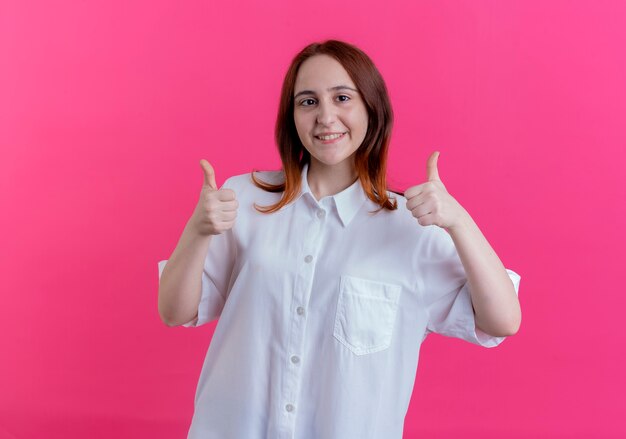 Sonriente joven pelirroja sus pulgares arriba aislados en rosa