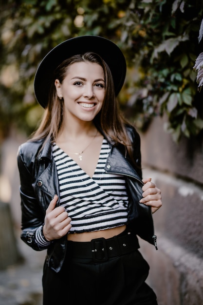 Sonriente joven de pelo castaño con una chaqueta de cuero, sombrero negro en el paseo de la ciudad