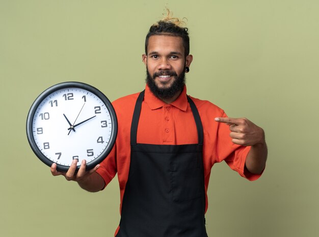 Sonriente joven peluquero afroamericano vistiendo uniforme sosteniendo apuntando al reloj