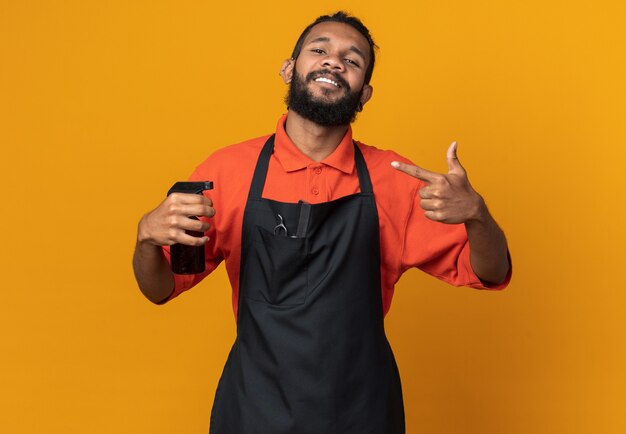 Sonriente joven peluquero afroamericano vistiendo uniforme sosteniendo y apuntando a laca para el cabello