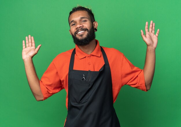 Sonriente joven peluquero masculino afroamericano vistiendo uniforme mostrando las manos vacías