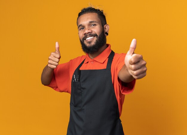 Sonriente joven peluquero masculino afroamericano vistiendo uniforme mostrando los pulgares para arriba