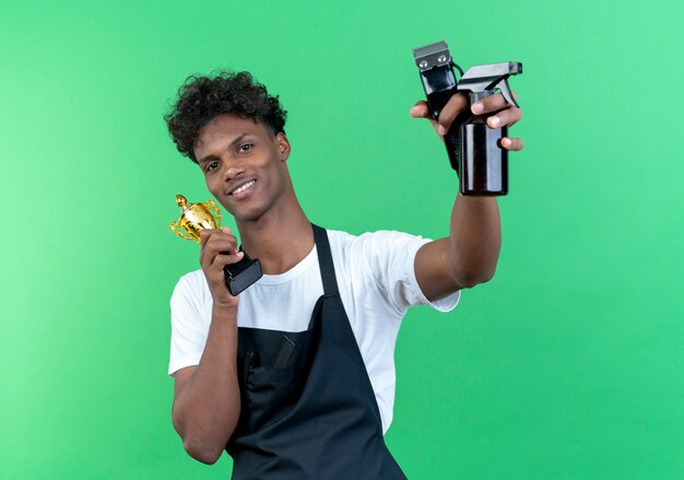 Sonriente joven peluquero masculino afroamericano vistiendo uniforme sosteniendo la copa ganadora alrededor de la cara y levantando herramientas de peluquero aisladas en la pared verde