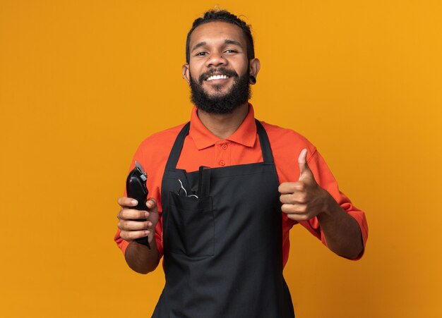 Sonriente joven peluquero masculino afroamericano vistiendo uniforme sosteniendo cortapelos mostrando el pulgar hacia arriba