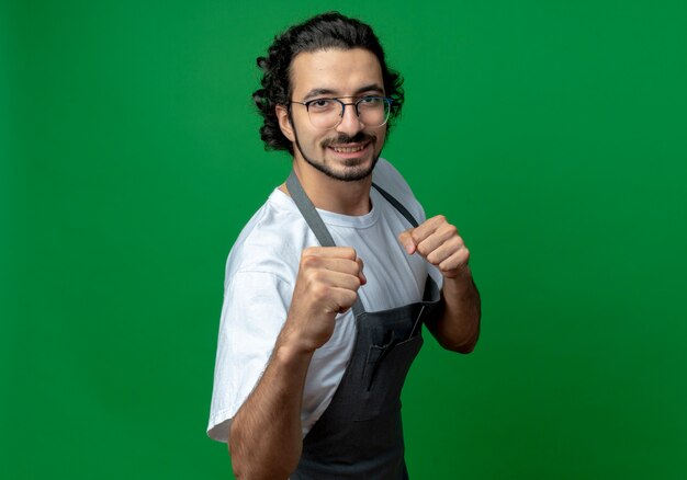 Sonriente joven peluquero masculino caucásico con gafas y banda de pelo ondulado en uniforme haciendo gesto de boxeo en cámara aislada sobre fondo verde con espacio de copia