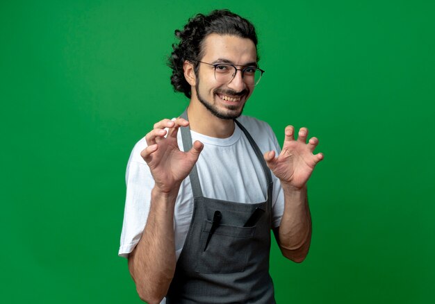 Sonriente joven peluquero masculino caucásico con gafas y banda de pelo ondulado en uniforme haciendo gesto de patas de tigre aislado sobre fondo verde con espacio de copia