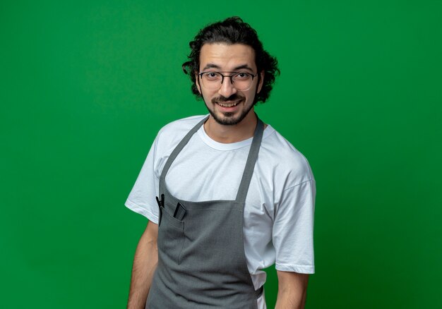 Foto gratuita sonriente joven peluquero masculino caucásico con gafas y banda para el pelo ondulado en uniforme de pie y mirando a cámara aislada sobre fondo verde con espacio de copia