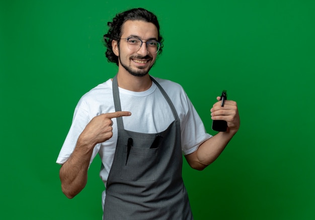 Sonriente joven peluquero masculino caucásico con gafas y banda de pelo ondulado en uniforme sosteniendo y apuntando a la botella de spray