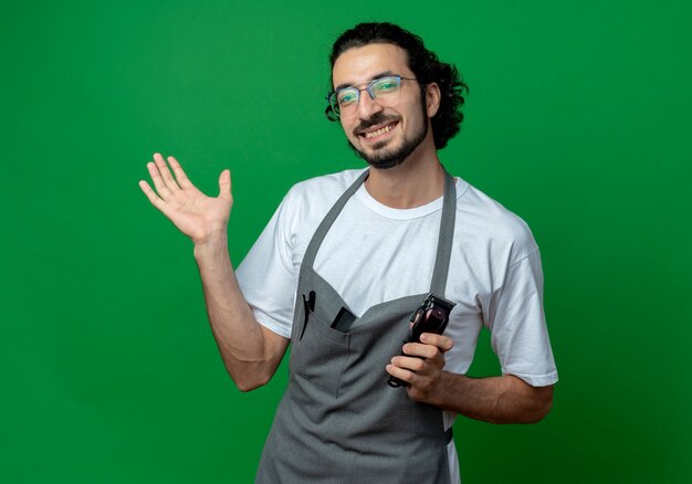 Sonriente joven peluquero masculino caucásico con gafas y banda para el pelo ondulado en uniforme sosteniendo cortapelos y mostrando la mano vacía aislada sobre fondo verde con espacio de copia