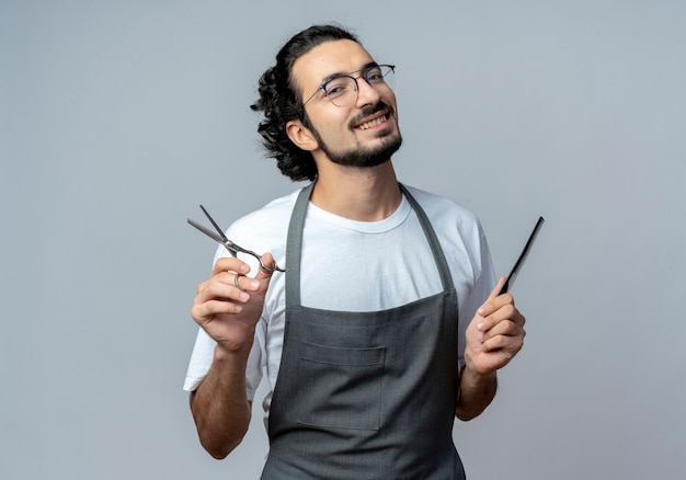 Foto gratuita sonriente joven peluquero masculino caucásico con gafas y banda para el pelo ondulado en uniforme sosteniendo tijeras y peine aislado sobre fondo blanco con espacio de copia