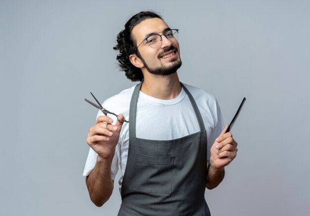 Sonriente joven peluquero masculino caucásico con gafas y banda para el pelo ondulado en uniforme sosteniendo tijeras y peine aislado sobre fondo blanco con espacio de copia