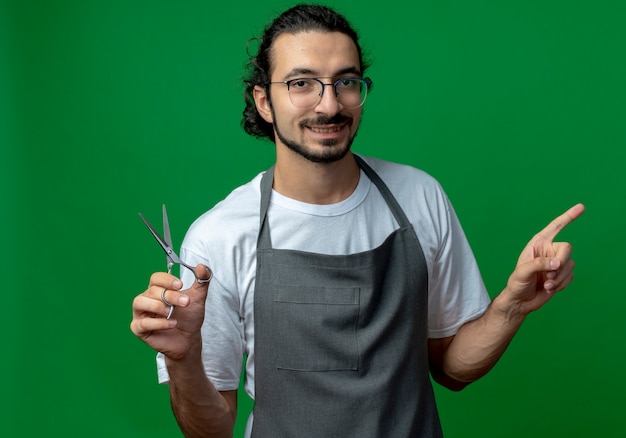 Sonriente joven peluquero masculino caucásico vistiendo uniforme y gafas sosteniendo tijeras y apuntando al lado aislado sobre fondo verde