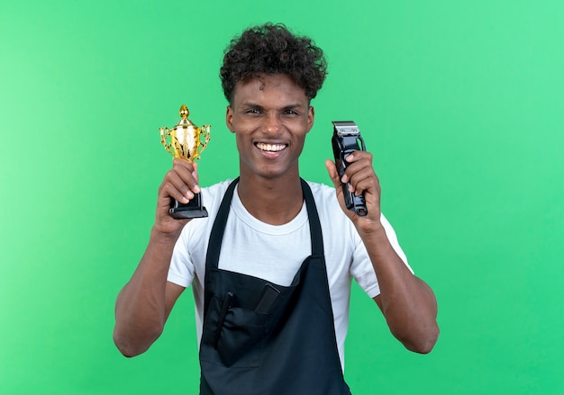 Sonriente joven peluquero vistiendo uniforme sosteniendo la copa ganadora con cabello