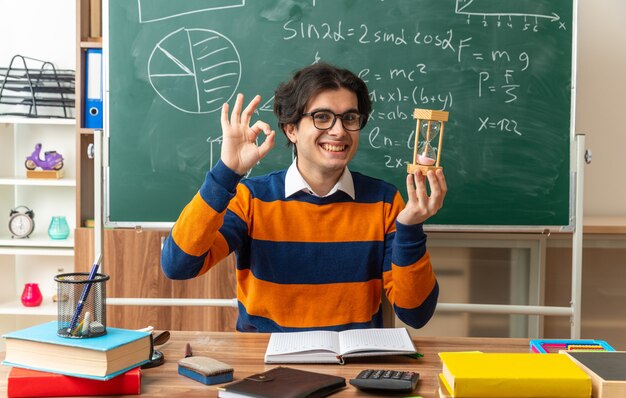 Sonriente joven profesor de geometría con gafas sentado en un escritorio con útiles escolares en el aula sosteniendo el reloj de arena mirando al frente haciendo bien firmar