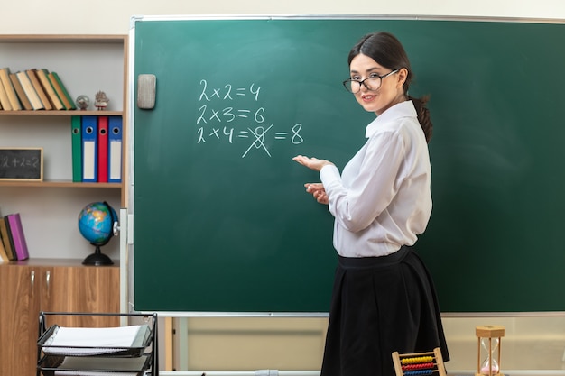 Sonriente joven profesora con gafas de pie detrás de la mesa con puntos de herramientas escolares con la mano en la pizarra en el aula