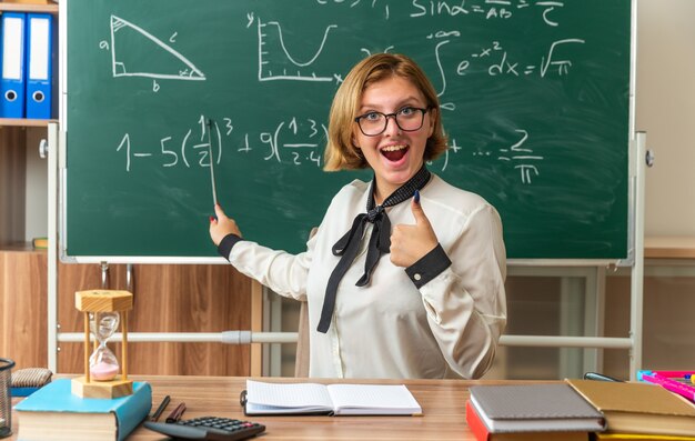 Sonriente joven profesora con gafas se sienta a la mesa con útiles escolares puntos en la pizarra con el puntero que muestra el pulgar hacia arriba en el aula