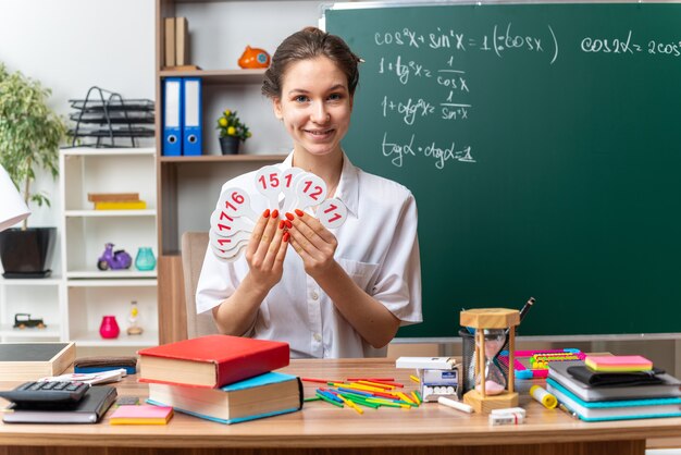 Sonriente joven profesora de matemáticas sentada en un escritorio con útiles escolares mirando al frente mostrando el número de ventiladores al frente en el aula