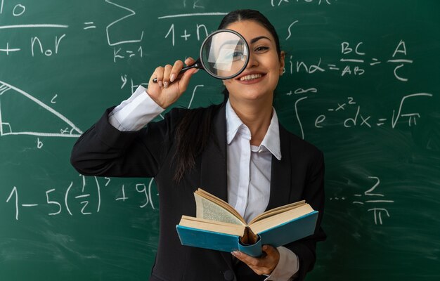 Sonriente joven profesora de pie delante de la pizarra mirando a la cámara con lupa sosteniendo el libro en el aula