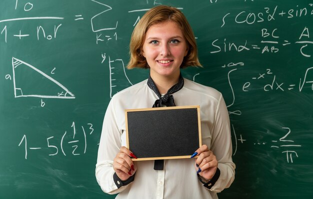 Sonriente joven profesora de pie delante de la pizarra sosteniendo una mini pizarra en el aula