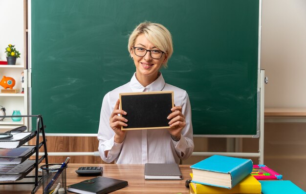 Sonriente joven profesora rubia con gafas sentado en un escritorio con herramientas escolares en el aula mirando a la cámara mostrando mini pizarra