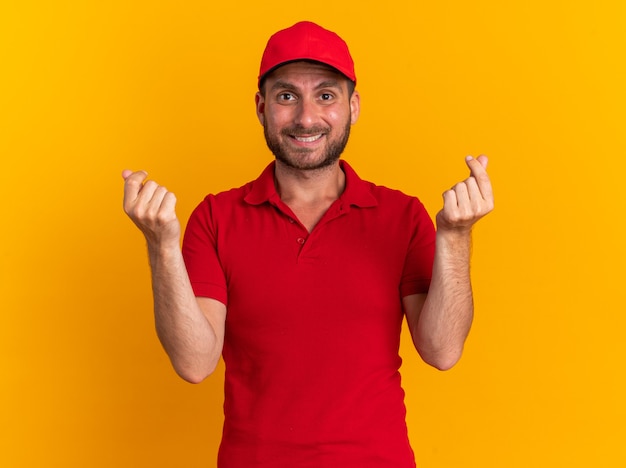 Sonriente joven repartidor caucásico en uniforme y gorra haciendo gesto de dinero