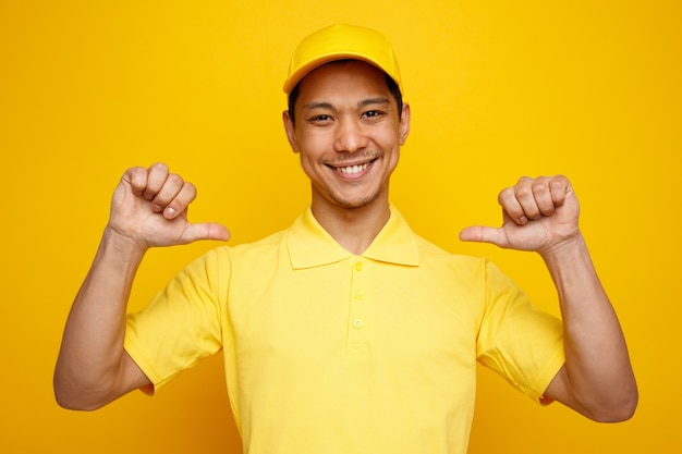 Sonriente joven repartidor vistiendo gorra y uniforme apuntando a sí mismo