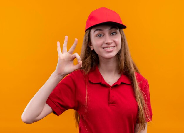 Sonriente joven repartidora en uniforme rojo haciendo bien firmar en espacio naranja aislado