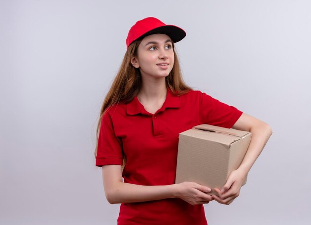 Sonriente joven repartidora en uniforme rojo sosteniendo la caja mirando hacia el lado derecho en un espacio en blanco aislado con espacio de copia