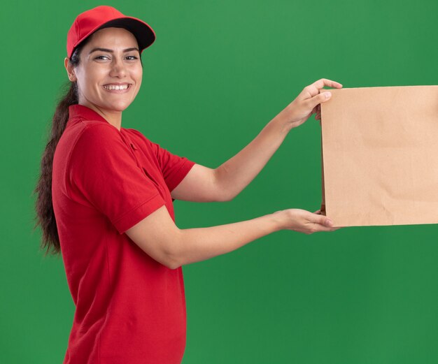 Sonriente joven repartidora vestida con uniforme y gorra dando paquete de comida de papel al cliente aislado en la pared verde