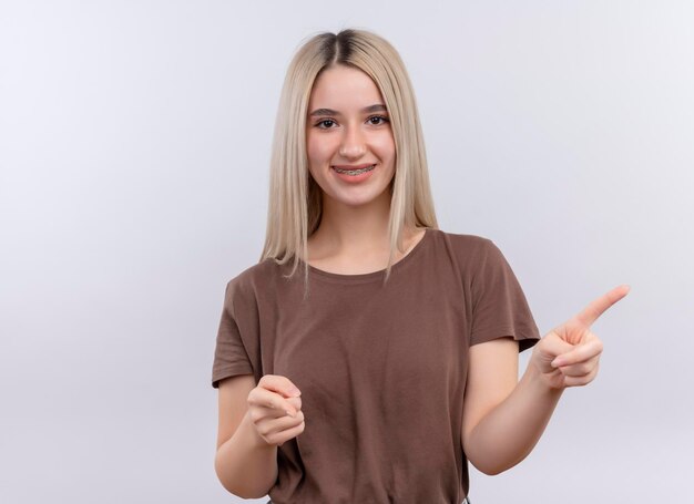 Sonriente joven rubia en aparatos dentales apuntando hacia el lado derecho en un espacio en blanco aislado con espacio de copia