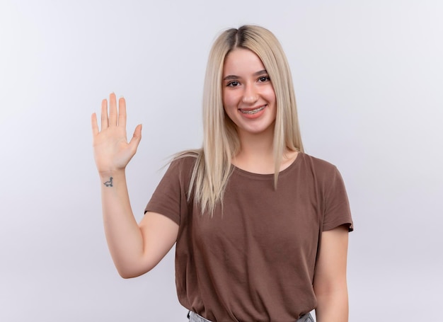 Sonriente joven rubia en aparatos dentales gesticulando Hola en espacio en blanco aislado