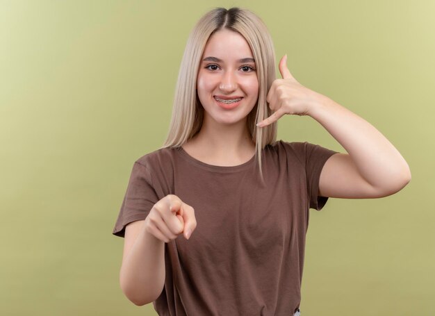 Sonriente joven rubia en aparatos dentales haciendo gesto de llamada y apuntando a un espacio verde aislado con espacio de copia