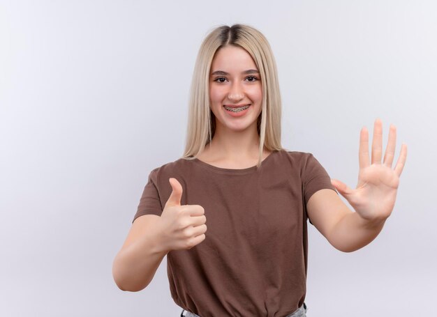 Sonriente joven rubia en aparatos dentales mostrando cinco y pulgar hacia arriba en un espacio en blanco aislado con espacio de copia