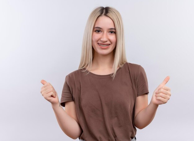 Sonriente joven rubia en aparatos dentales mostrando los pulgares para arriba en el espacio en blanco aislado