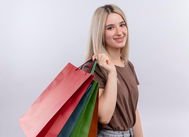 Sonriente joven rubia en aparatos dentales sosteniendo bolsas de compras en el hombro en un espacio en blanco aislado