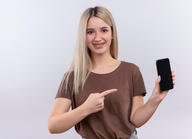 Sonriente joven rubia en aparatos dentales sosteniendo el teléfono móvil y apuntando a él en un espacio en blanco aislado con espacio de copia