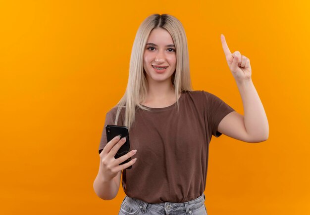 Sonriente joven rubia en aparatos dentales sosteniendo el teléfono móvil con el dedo levantado en el espacio naranja aislado con espacio de copia