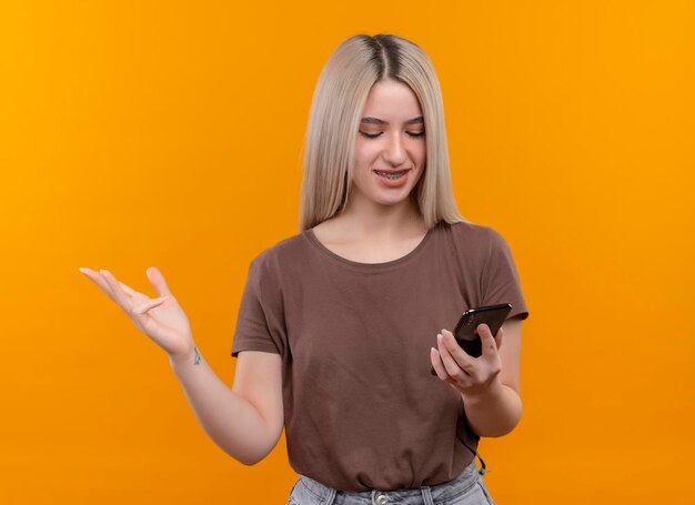 Sonriente joven rubia en aparatos dentales sosteniendo teléfono móvil mirándolo mostrando la mano vacía en el espacio naranja aislado con espacio de copia