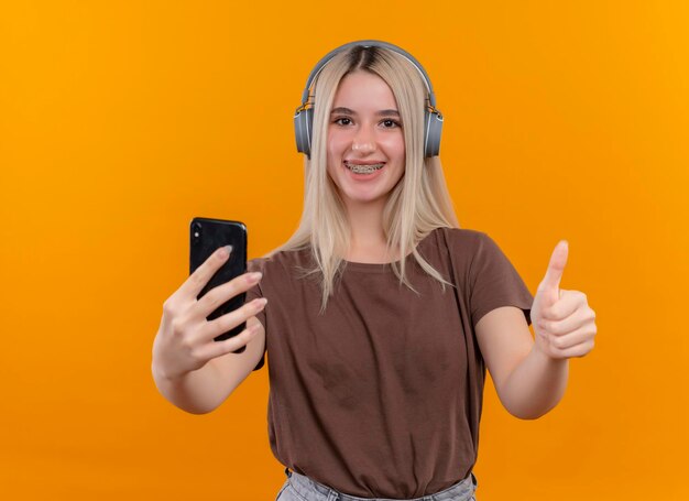 Sonriente joven rubia con audífonos en aparatos dentales sosteniendo el teléfono móvil y mostrando el pulgar hacia arriba en el espacio naranja aislado