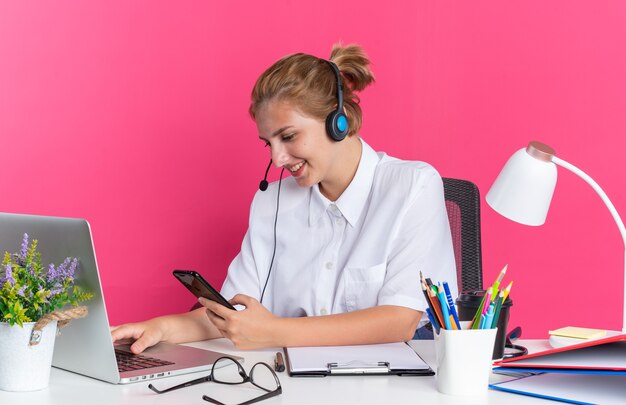 Sonriente a joven rubia centro de llamadas chica con auriculares sentado en el escritorio con herramientas de trabajo usando una computadora portátil y un teléfono móvil aislado en la pared rosa