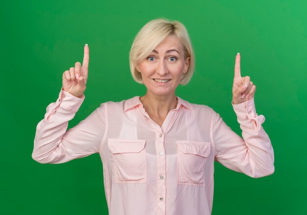 Sonriente joven rubia eslava apuntando hacia arriba aislado sobre fondo verde