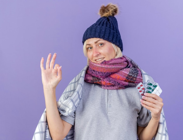 Sonriente joven rubia eslava enferma vistiendo gorro de invierno y bufanda envuelta en plaid gestos signo de mano ok