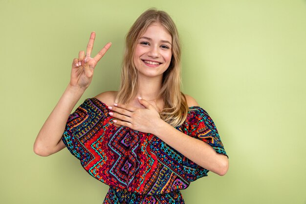 Sonriente joven rubia eslava poniendo la mano sobre su pecho y gesticulando el signo de la victoria